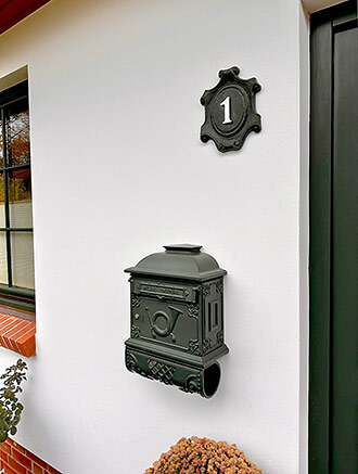Wandbriefkasten mit Zeitungsrolle (Art.-Nr. 1524 AZ) in Tannengrün mit einer individuell gegossenen Einwurfklappe. Darüber hängt ein passendes Hausnummernschild (Art.-Nr. 403) mit weiß lackierter Hausnummernzahl.